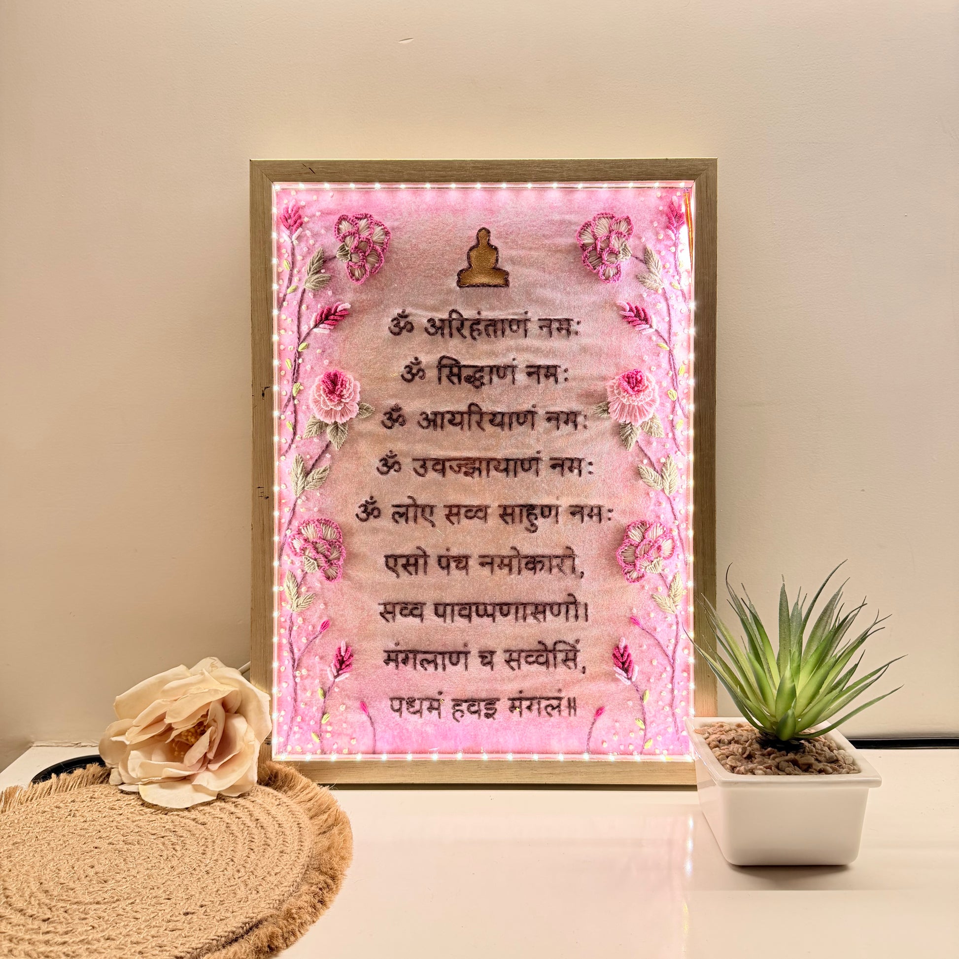 Decorative framed artwork with pink floral design and text on a table with a plant and flower and navkar mantra 
